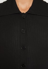 Närbild av ett svart kabelstickat plagg med fyra stora svarta knappar och en bred krage, bärs på en persons övre bröst.