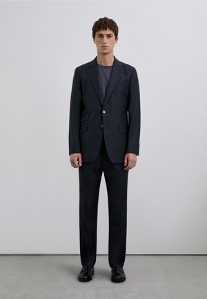 Traje oscuro a medida con blazer estructurado y pantalones a juego, que cuenta con un botón único y dos bolsillos frontales, combinado con una camiseta gris.