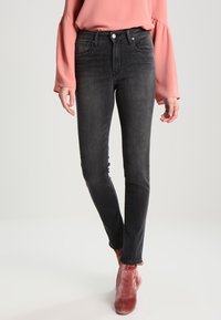 Femme portant un jean skinny noir délavé, un haut ample à manches longues rose, et des bottines en velours rougeâtre, debout devant un fond uni.
