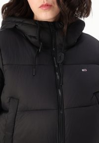 Czarna kurtka puchowa z kapturem, wyposażona w zamek błyskawiczny z przodu, regulowane sznurki oraz małe logo po lewej stronie klatki piersiowej. Teksturowana powierzchnia.