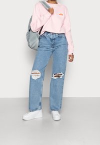 Felpa rosa chiaro con un piccolo logo, indossata con jeans blu a vita alta con grandi strappi sulle ginocchia e sneakers bianche con zeppa.