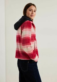 Red and white plaid jacket with a black, lightweight hood. Features long sleeves and pockets. Worn with dark blue jeans. Smooth texture.
