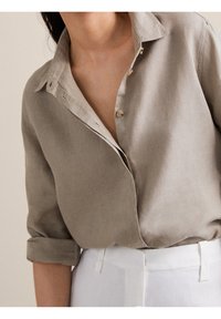 Chemise en lin taupe clair avec col et petits boutons, manches retroussées et coupe décontractée. Associée à un pantalon blanc taille haute.