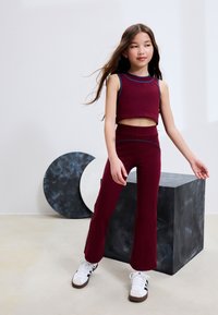 Top corto color mogano e pantaloni a zampa a vita alta, con accenti blu, realizzati in materiale elasticizzato. Modella in piedi su un piedistallo geometrico.