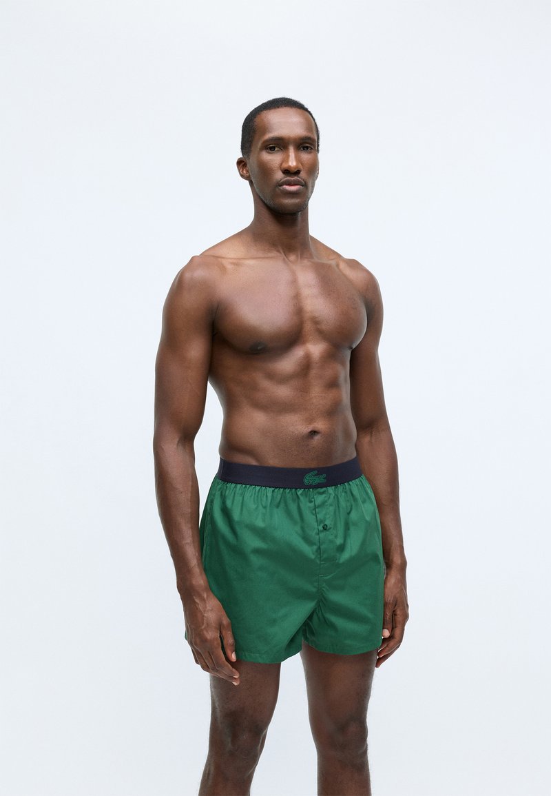 Green swimming shorts made of lightweight material, featuring a dark waistband with a logo, button front, and relaxed fit design.