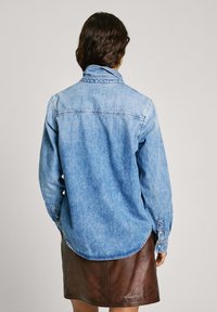 Svetlo modra denim srajca z ovratnikom na gumb, dolgimi rokavi in okroglim robom. Ima vidne šive in klasične manšete.