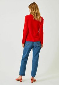 Punainen pitkähihaiseksi neuleeksi yhdistetty sinisiin suoriin farkkuihin ja oransseihin korkokenkiin. Neuleessa on istuva muotoilu ja sileä tekstuuri.