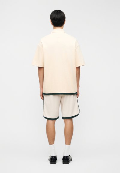 Άνδρας όρθιος γυρισμένος πλαγίως, φοράει κρεμ κοντομάνικο πουκάμισο και ταιριαστά σορτς με σκούρες λεπτομέρειες, λευκές κάλτσες και μαύρα επίσημα παπούτσια.