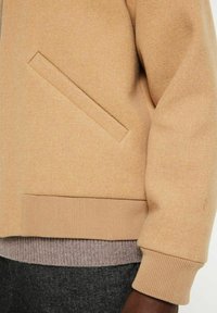 Beige Jacke mit gerippten Bündchen und Saum, ausgestattet mit einer Seitentasche, aus weichem, strukturiertem Stoff, über einem grauen Strickkleidungsstück getragen.