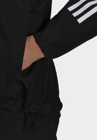 Giacca nera con maniche lunghe, caratterizzata da dettagli striati bianchi sulle maniche. Include una vita arricciata e una texture liscia e leggera.