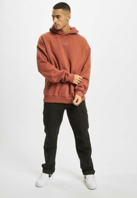 Haut à capuche rouge brique avec logo "DEF", coupe oversize, poignets et ourlet côtelés, associé à un pantalon cargo noir et des baskets blanches.