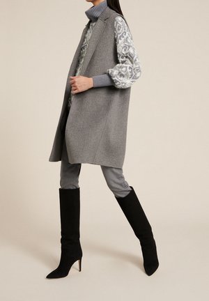 Frau trägt einen grauen ärmellosen Mantel über einem gemusterten Pullover, graue Hose und schwarze hochhackige kniehohe Stiefel, die vor einem beigen Hintergrund läuft.