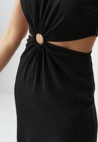 Robe noire avec un design tordu et une découpe circulaire à la taille. Fabriquée en tissu doux avec une texture froncée autour de la découpe.