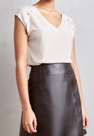 Kobieta ubrana w beżowy, bezrękawnik z dekoltem w kształcie litery V z srebrnymi guzikami oraz wysoką, czarną skórzaną spódnicę na tle jednolitego tła.