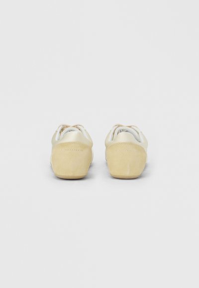 Ζευγάρι μπεζ σουέτ και δερμάτινα αθλητικά παπούτσια, σε προβολή από πίσω, τοποθετημένα σε λευκή επιφάνεια με τα κορδόνια χαλαρά δεμένα.