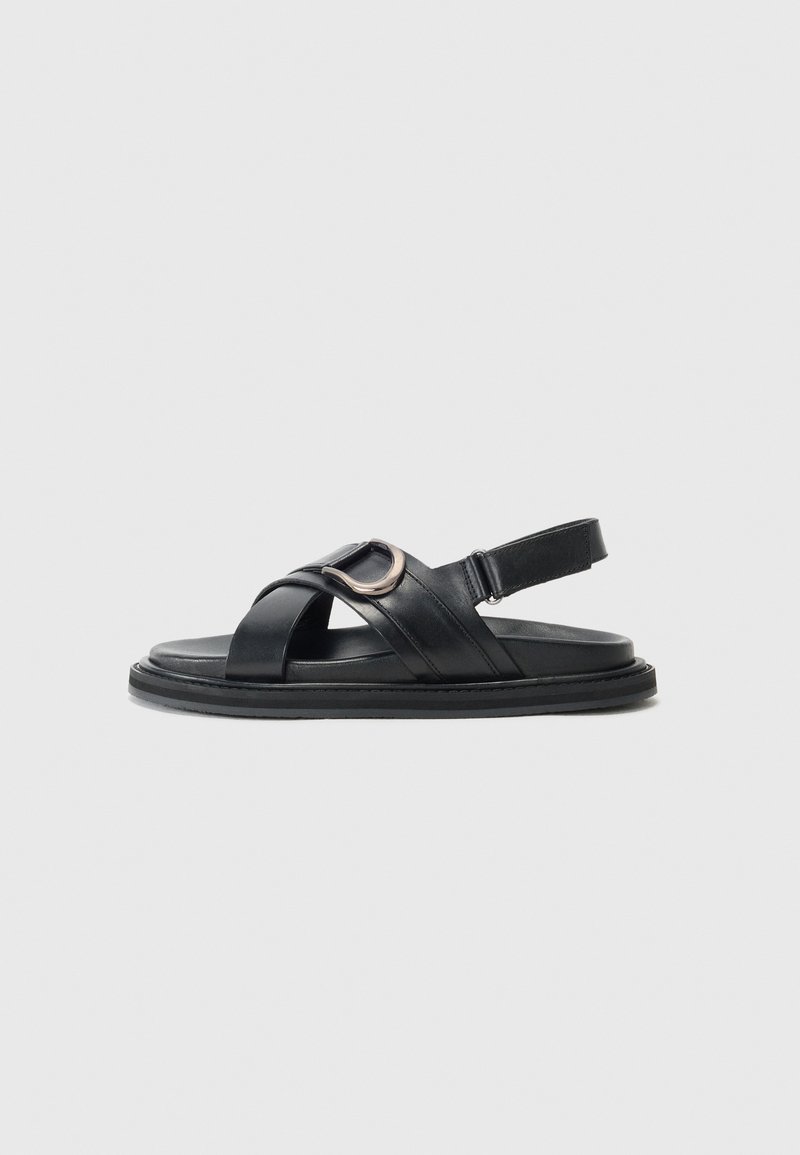 Black leather sandal with crisscross straps, a circular metal accent, and adjustable back strap. Flat sole with a textured finish.