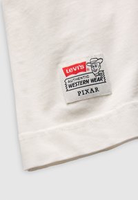 Bílá bavlněná tkanina s cedulkou Levi's, na které je ilustrace kovboje a text "AUTENTICKÉ ZÁPADNÍ OBLEČENÍ PIXAR."