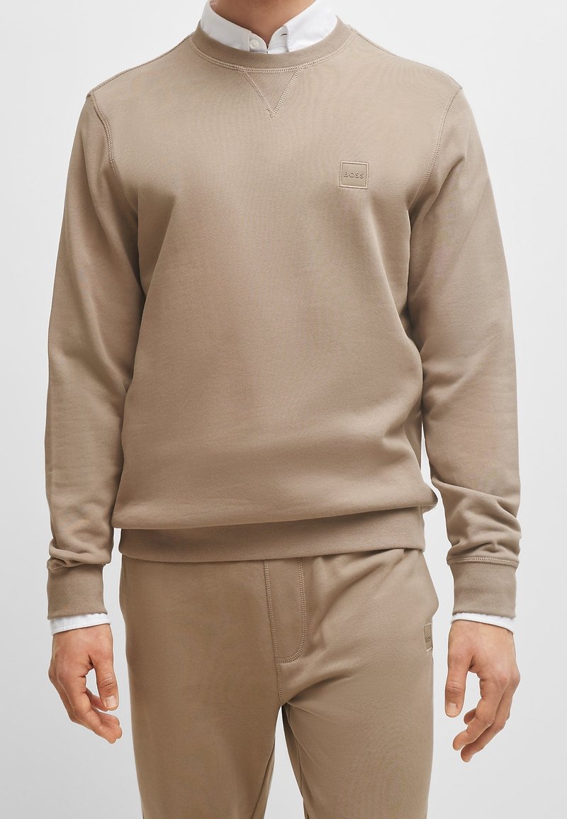 Homme portant un sweat-shirt beige et un pantalon assorti avec un petit logo sur la poitrine, col de chemise blanc visible sous l'encolure du sweat-shirt.