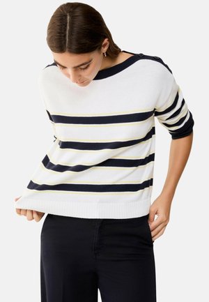 Donna con un maglione bianco con righe orizzontali blu navy e gialle, che sistema l'orlo, abbinato a pantaloni neri.