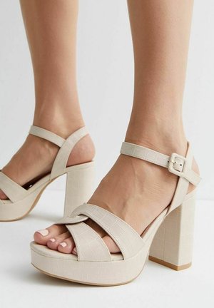 Kvinde iført beige højhælede sandaler med plateau såler og ankelremme med spænde, der viser tæerne og en del af underbenene.