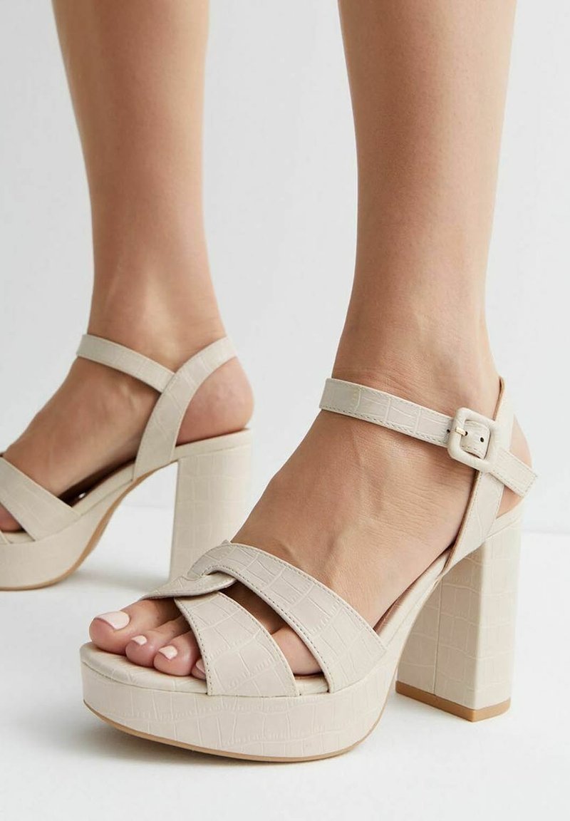 Femme portant des sandales beige à talons hauts avec semelles compensées et brides à la cheville avec boucle, montrant les orteils et une partie des jambes inférieures.