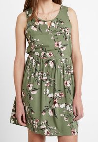 Vestido floral en verde oliva con un cuerpo ajustado, escote de ojal y cintura elástica. Presenta un patrón de flores en rosa claro y blanco.