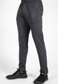 Grijze sportbroeken met een elastische tailleband, een zijzak met ritssluiting en een blauw logo. De stof lijkt glad met een lichte glans.