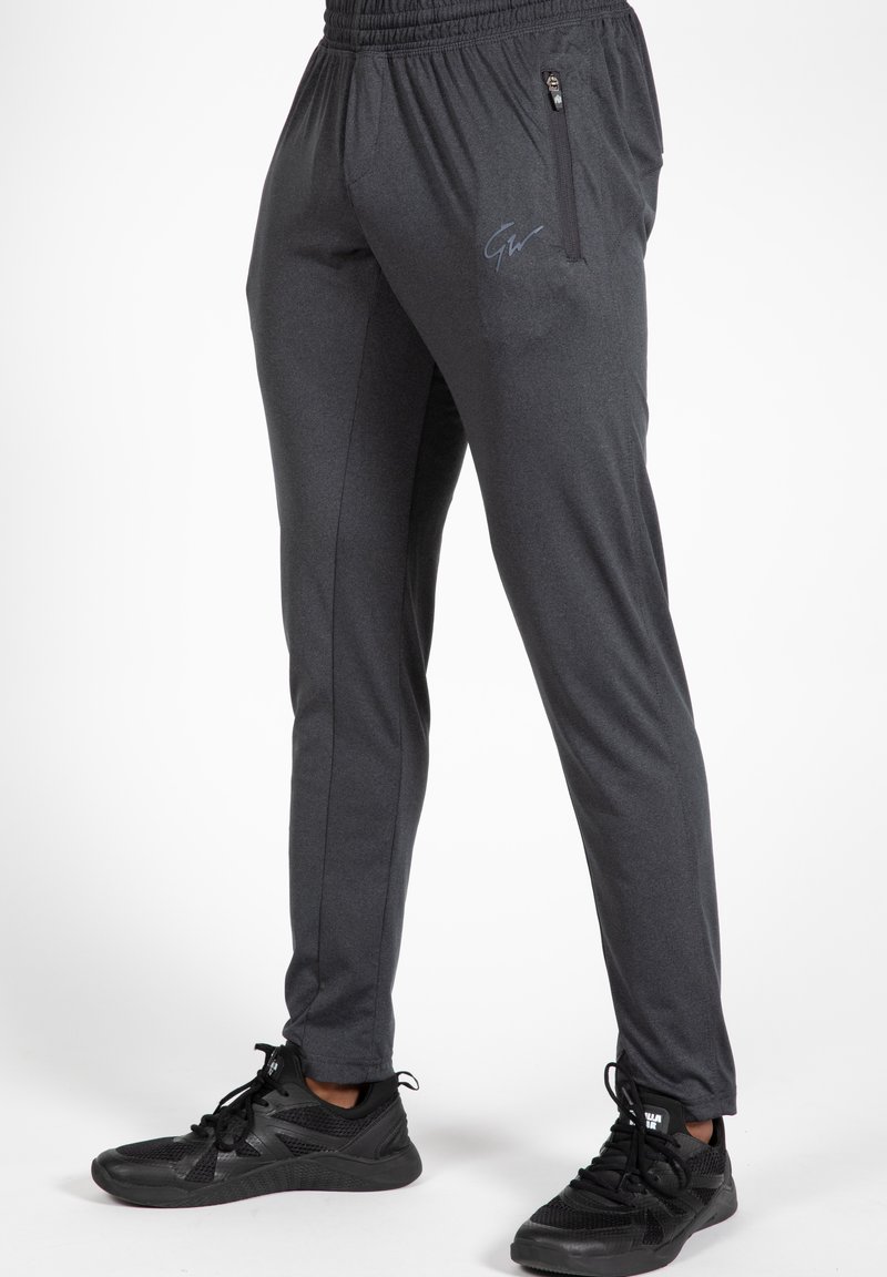 Grijze sportbroeken met een elastische tailleband, een zijzak met ritssluiting en een blauw logo. De stof lijkt glad met een lichte glans.