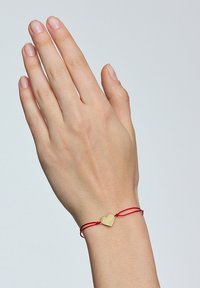 Czerwona bransoletka z nicią z złotym zawieszką w kształcie serca, prosta konstrukcja, gładka tekstura i regulowane zapięcie, noszona na dłoni z naturalnymi paznokciami.