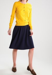 Gul stickad kofta med rund halsringning och knappar, kombinerad med en mörk marinblå kjol med utsvängd skärning och bruna loafers med leopardmönster.