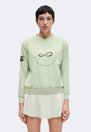 Ung person med kort mørkt hår bærer lysegrøn sweatshirt med uendelighedssymbol og tekst samt hvid tekstureret nederdel.
