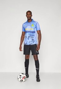 Muž v modrom futbalovom dresu, čiernych šortkách a ponožkách stojí s jednou nohou na futbalovej lopte na jednofarebnom pozadí.