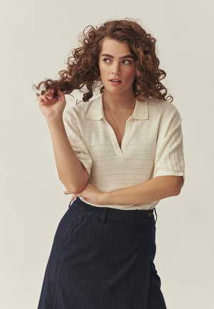 Jeune femme aux cheveux bouclés portant un haut crème côtelé à manches courtes et une jupe taille haute foncée, tenant une boucle de cheveux et regardant sur le côté.