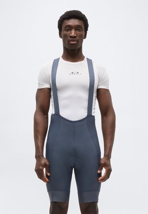 Atlétikus férfi fehér Oakley rövid ujjú felsőben és sötétszürke, vállpántos kerékpáros nadrágban, sima háttér előtt állva.