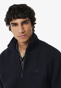 Pullover in pile di colore blu navy con zip a un quarto, colletto alto e logo ricamato. Tessuto strutturato e strato visibile sotto.