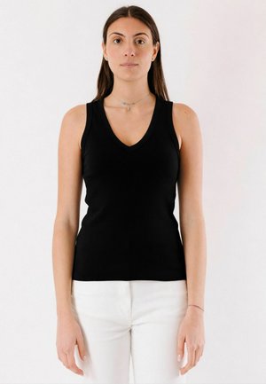 Giovane donna con capelli castani lunghi, che indossa un top nero senza maniche con scollo a V e pantaloni bianchi, in piedi davanti a uno sfondo chiaro e neutro.