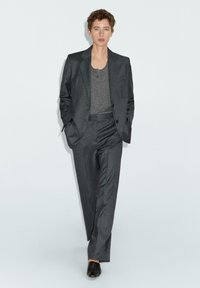 Grau maßgeschneideter Anzug mit einem taillierten Blazer und geraden Hosen, kombiniert mit einem dunkelgrauen Oberteil und schwarzem Schuhwerk. Schlichter, strukturierter Stoff.