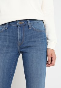 Blå denimjeans med en slät yta, med knappstängning, femfickorsdesign och kontrasterande sömmar. Något figursydd vid midjan.