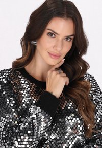 Schwarzer Pullover mit silbernen Pailletten, rundem Halsausschnitt und engen Bündchen. Das Model hat lange, gewellte Haare und einen Ring an ihrem Finger.