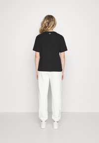 Frau mit welligem blonden Haar steht vor einer weißen Wand, trägt ein schwarzes T-Shirt mit kurzen Ärmeln, weiße Jogginghosen mit Schriftzug an den Knöcheln und weiße Sneakers.