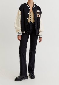 Černá a krémová varsity bunda se bílými pruhy, vyšitým znakem, nošená přes béžový žebrovaný top a černé džíny s vysokým pasem, doplněná o robustní boty.