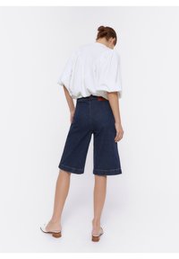 Mujer de pie de espaldas vistiendo una blusa blanca de mangas abullonadas, pantalones cortos de mezclilla oscuros hasta la rodilla y zapatos de tacón blanco sin cordones sobre un fondo blanco.