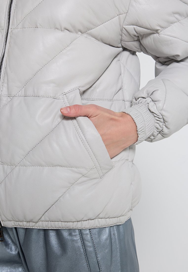 Chaqueta de cuero acolchada de color gris claro, con cierre de cremallera, puños elásticos y bolsillos laterales. Textura suave y patrón de costura geométrica.