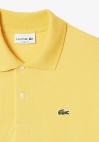 Polo amarillo hecho de una tela texturizada, con cuello tipo polo, tres botones y un logo de cocodrilo verde en el pecho.