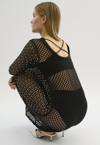 Rochie din plasă neagră cu o croială lejeră, având deschideri mari în formă de diamant și bretele subțiri la spate, purtată peste un strat de bază solid.
