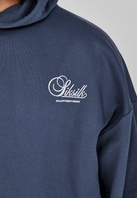 Tamno plavi dukserica od mekog materijala s kapuljačom, s bijelim izvezenim tekstom "Siksilk" i "FOLLOWTHEMOVEMENT" na prsima.