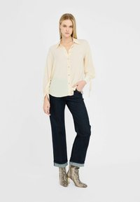 Cremefarbige Bluse mit durchgehender Knopfleiste und lockeren Ärmeln, kombiniert mit dunkler Denim-Jeans und gemusterten Absatzstiefeln.