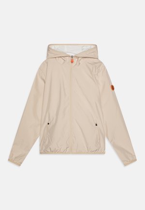 Giacca a vento beige con cappuccio, zip frontale, due tasche laterali, polsini elastici e una patch con logo sulla manica sinistra. Materiale liscio.