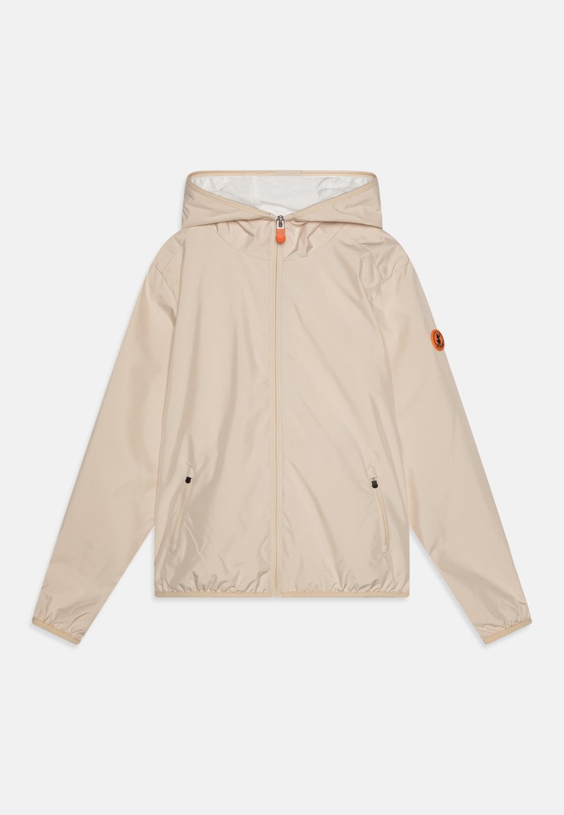 Veste coupe-vent beige avec capuche, fermeture éclair sur le devant, deux poches latérales, poignets élastiques et un patch logo sur la manche gauche. Matière lisse.