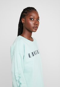 Sweatshirt verde menta com detalhes de mangas com folhos e um logo preto impresso "UPGREAT". Tecido suave e corte descontraído.